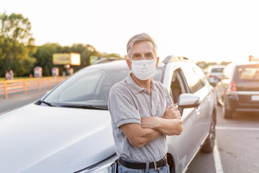 Older man wearing a facemask posing in front of his car